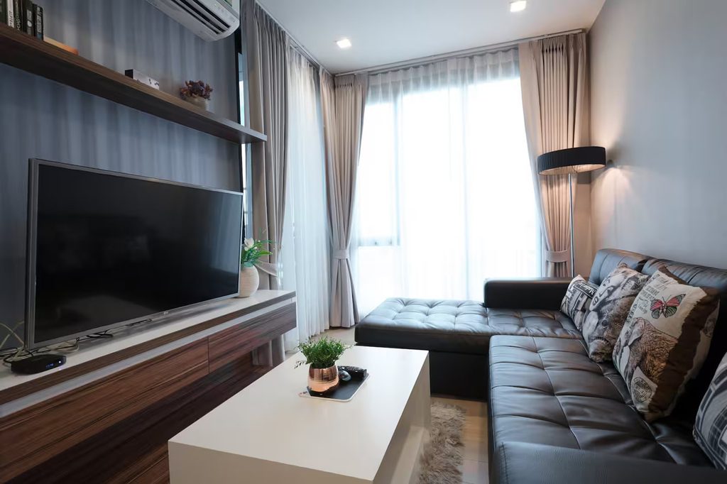 Furnished condo living room with an L-shaped sofa, television, coffee table, and large curtained windows