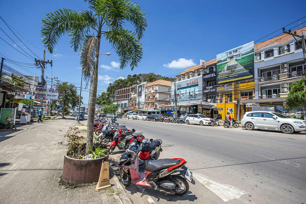 Street in Thailand with shops, low-rise buildings, palm trees, and parked motorbikes