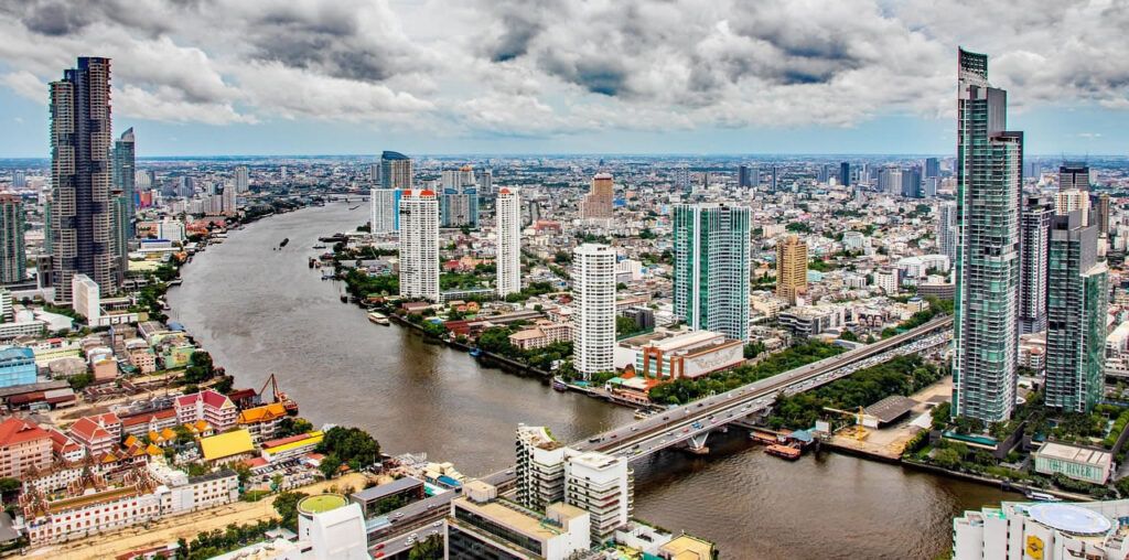 A high-angle view of downtown Bangkok featuring the Chao Phraya River, modern high-rises, and city infrastructure under a cloudy sky.