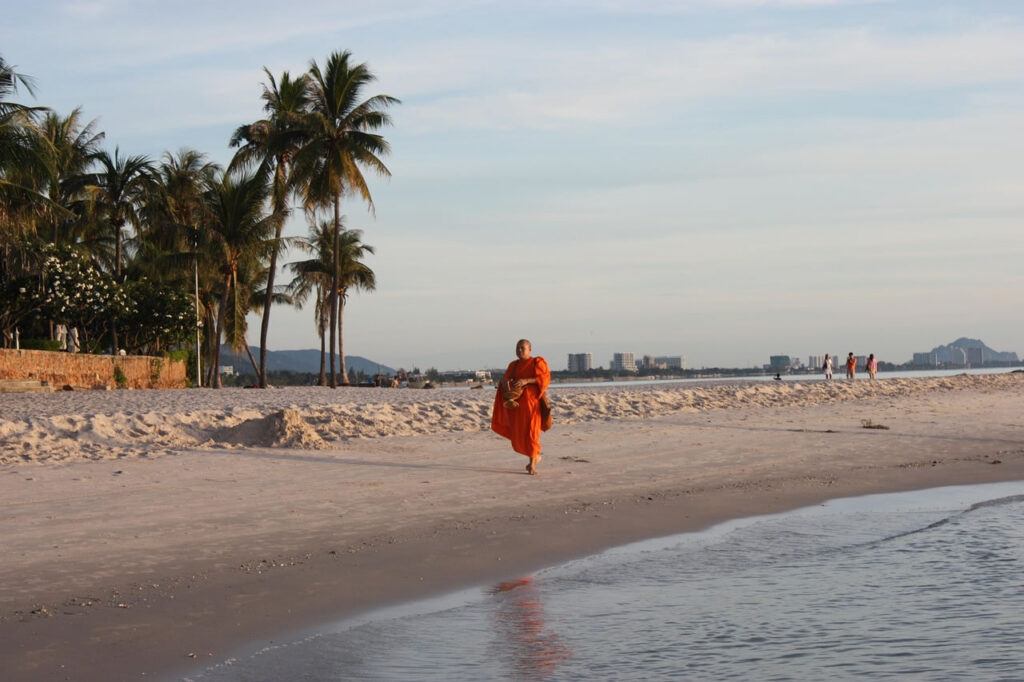 A Thai Buddhist monk walking barefoot along Hua Hin beach at sunrise, with palm trees, calm water, and a distant city skyline in the background.
