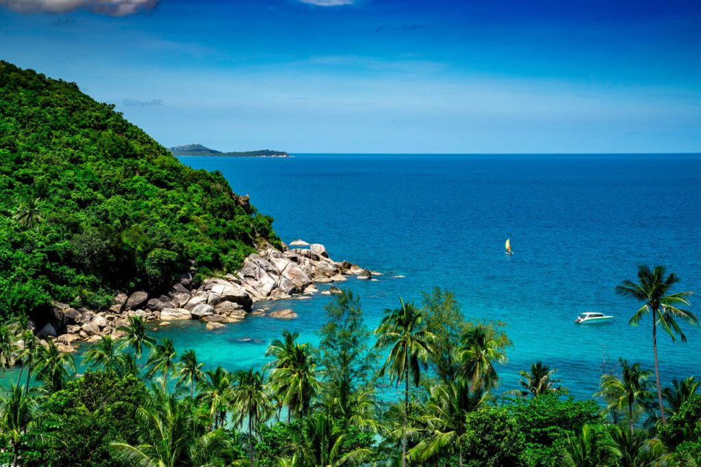 Tropical coastline view of Koh Samui, Thailand, with lush green hills, turquoise waters, palm trees, and boats dotting the calm sea.