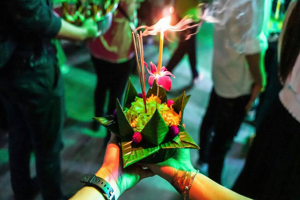 A person holds a handmade krathong decorated with flowers, banana leaves, incense, and a candle during a Loi Krathong celebration in Thailand.