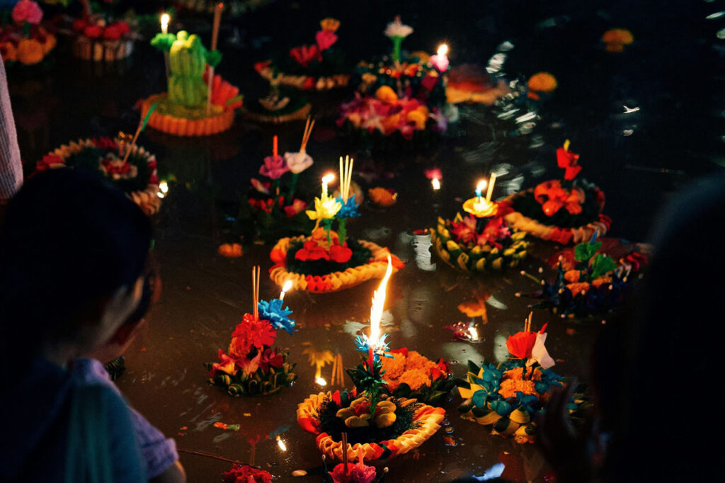 Colorful krathongs made of banana leaves, flowers, and candles float on a river during Thailand’s Loi Krathong festival, as people look on.