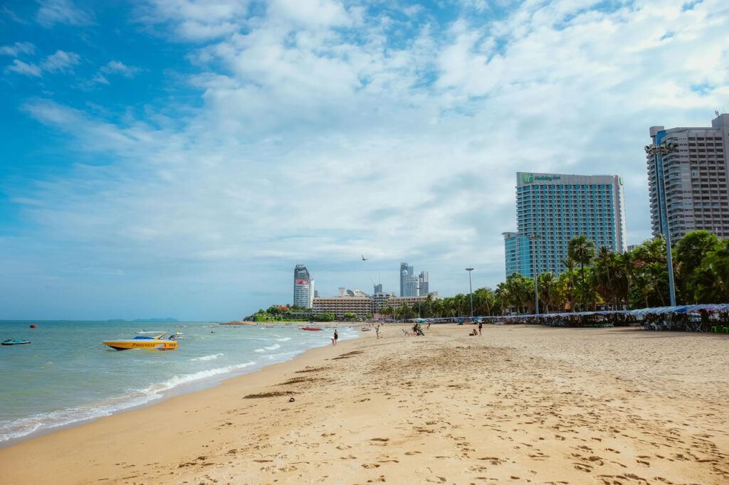 View of Pattaya Beach in Thailand with a yellow speedboat, high-rise buildings, palm trees, and beachgoers on a partly cloudy day.