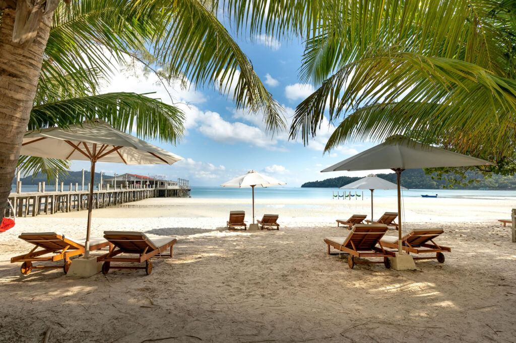Scenic view of a beach in Phuket, Thailand, with wooden sun loungers, umbrellas, palm trees, and a pier extending into calm turquoise waters.