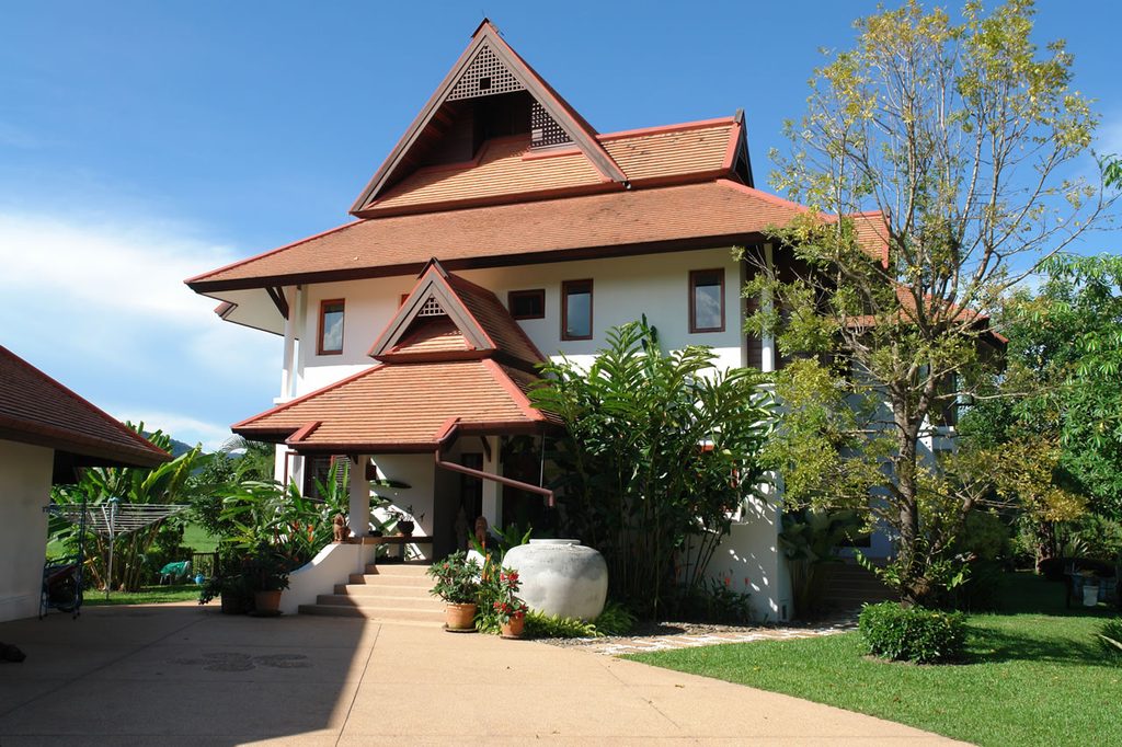 Two-storey Thai-style house with a red tiled roof, tropical garden, and paved driveway.