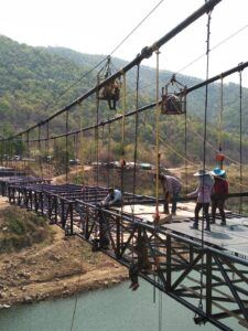 Workers constructing a suspended bridge over a river in rural Thailand, with some suspended in harnesses above and below the structure.
