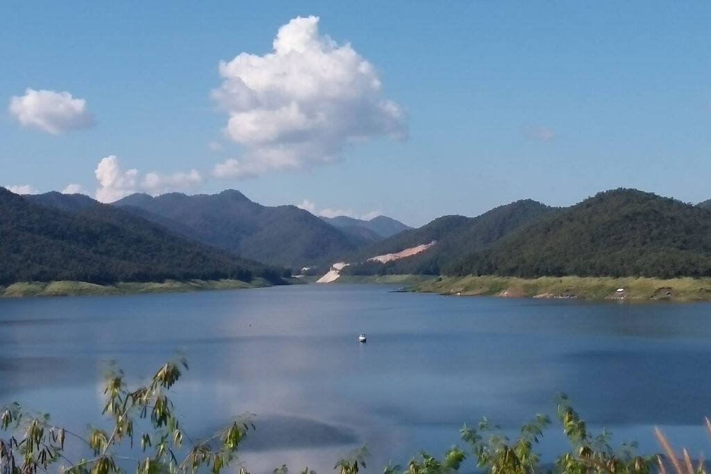 Peaceful view of Mae Kuang Lake near Chiang Mai, with calm blue water, forested mountains, and a boat in the distance under a bright sky.