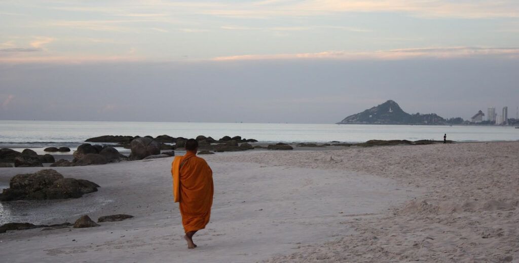 Buddhist monk walking along Hua Hin Beach with view of Khao Takiap hill in the background