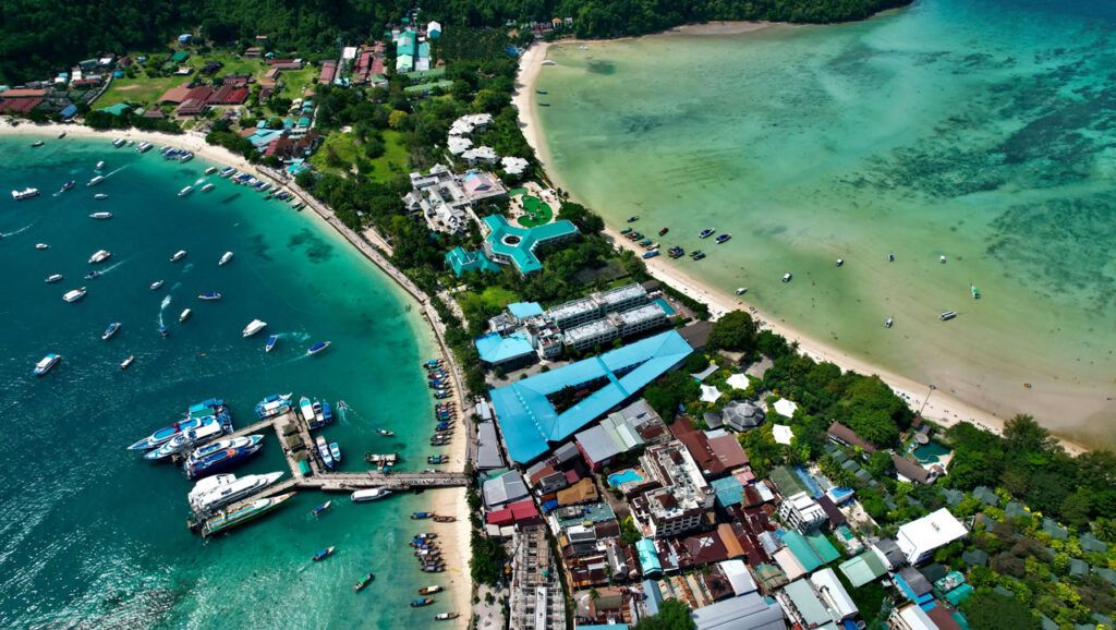 Aerial view of Phi Phi Don island showing the main pier, turquoise waters, beachfront buildings, and boats docked along the coast in Krabi, Thailand