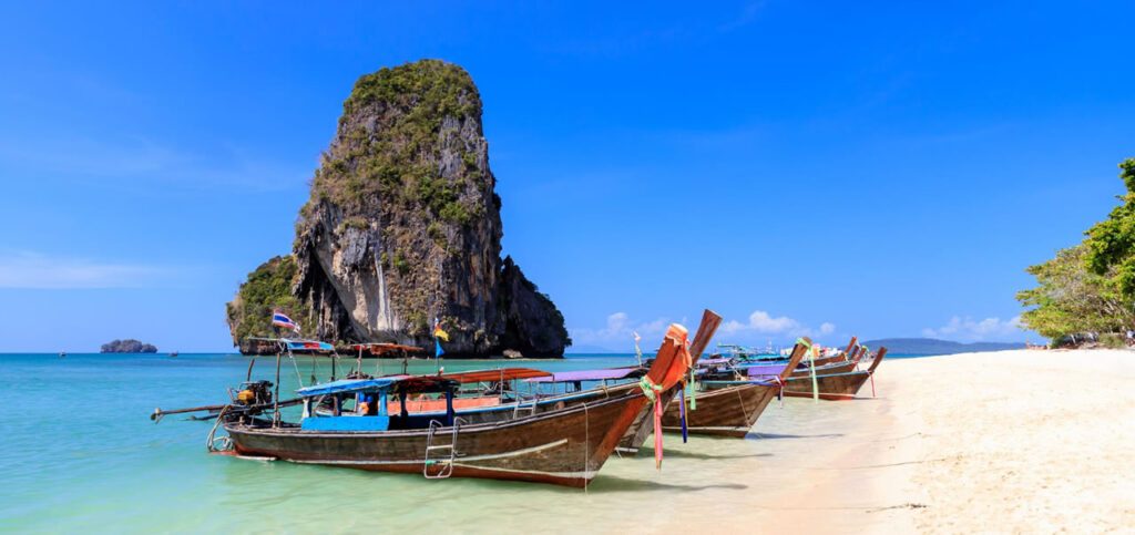 Traditional longtail boats moored on the white sands of Railay Beach in Krabi, Thailand with limestone cliffs and turquoise sea under a clear blue sky
