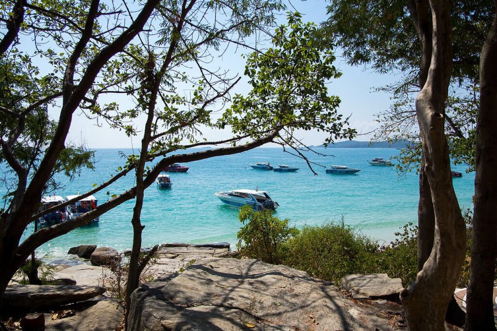 View of speedboats anchored in turquoise water near Koh Samet, framed by trees and rocky shoreline in the foreground