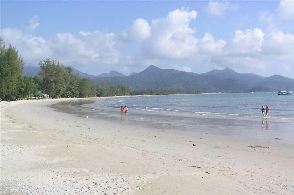 Klong Prao Beach on Koh Chang with a wide stretch of soft sand, calm blue waters, a few people strolling along the shore, and lush green mountains in the background under a partly cloudy sky.
