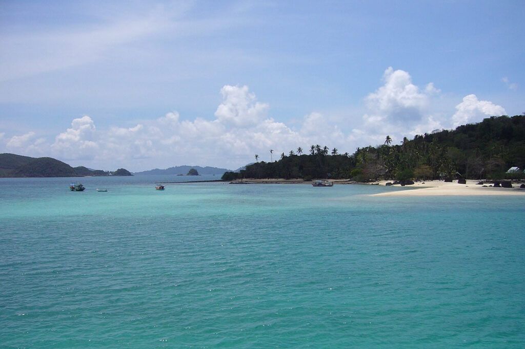 Panoramic view of southern Koh Chang with turquoise waters, sandy beach, palm trees, and fishing boats under a bright blue sky