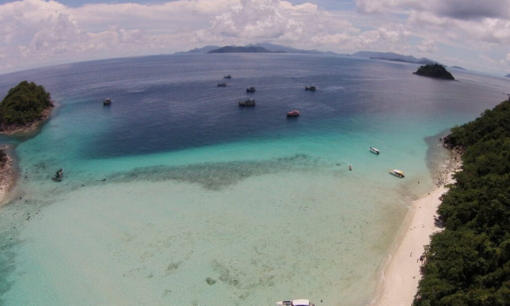 Aerial view from Koh Rang archipelago near Koh Chang, showing turquoise waters, coral reefs, anchored boats, and a small white-sand beach bordered by lush green forest.