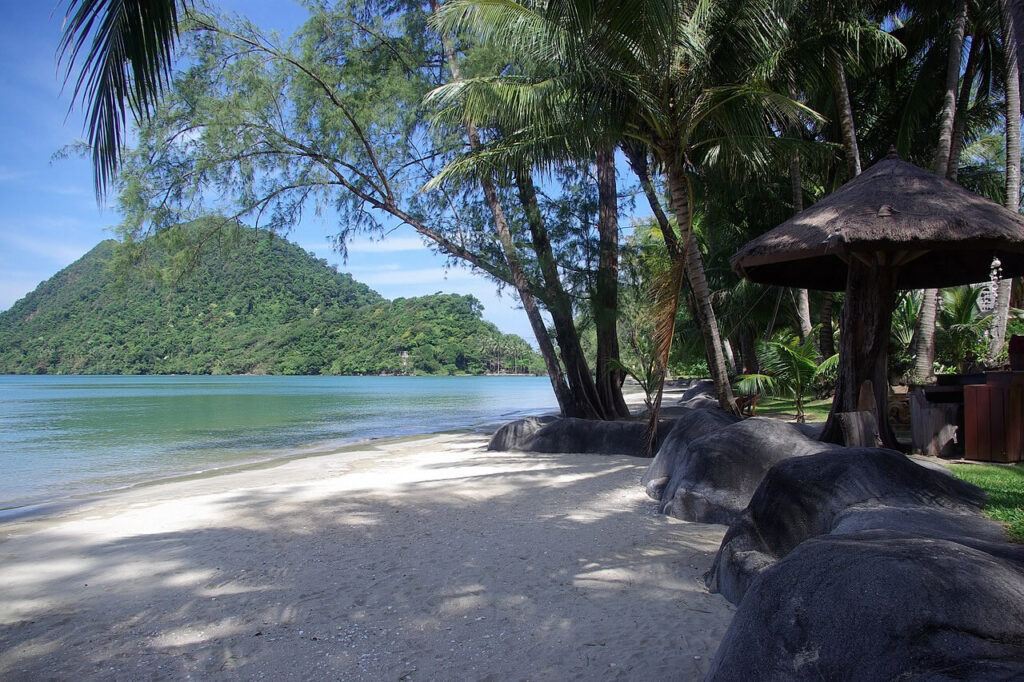 Secluded bay on Koh Chang with calm turquoise water, lush green hillside, palm trees, and shaded beach area.