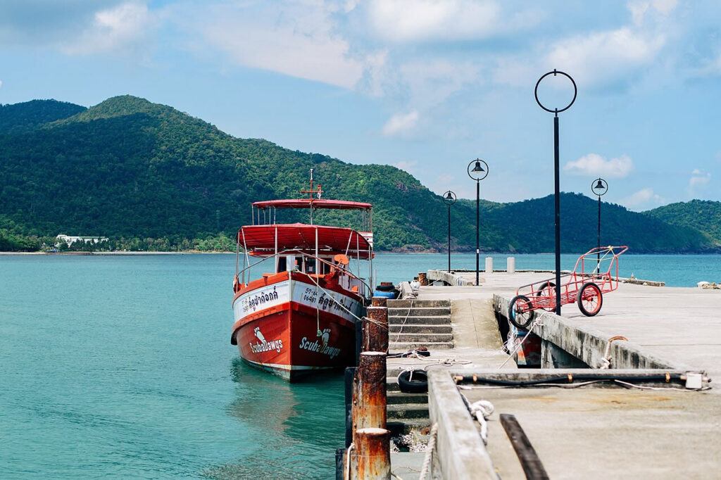 Red private boat docked at a pier with turquoise waters and lush green hills in the background on Koh Chang, Thailand