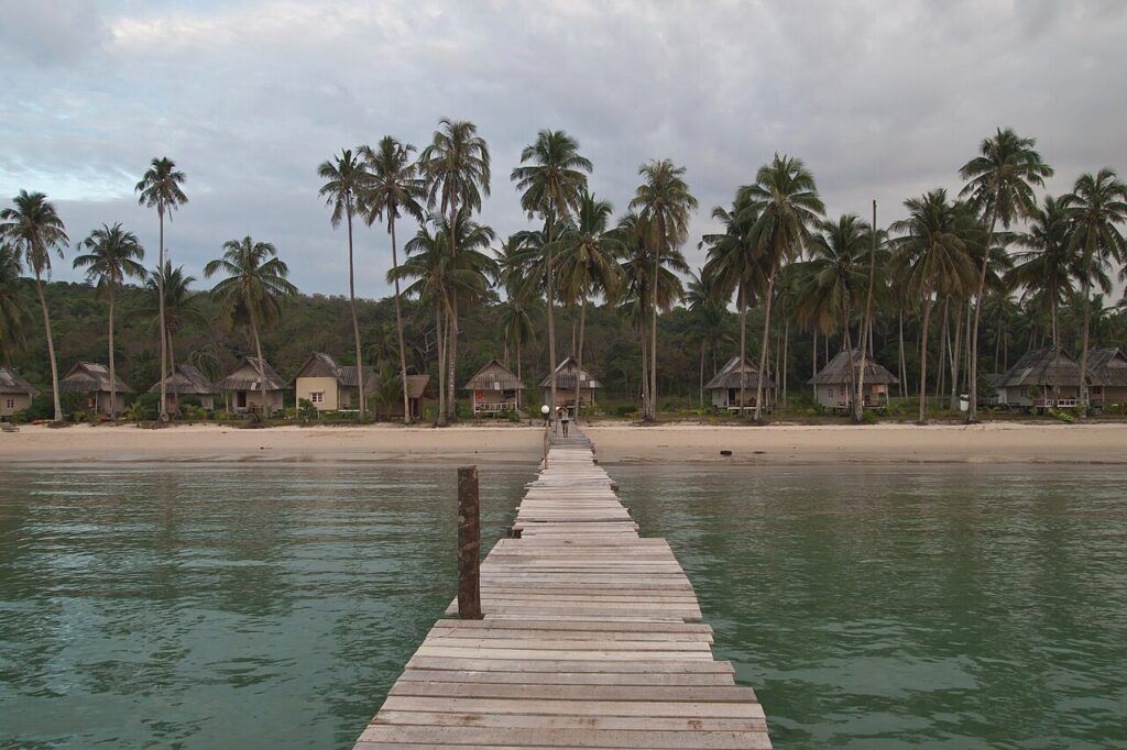 Wooden pier leading to Bang Bao Beach on Koh Kood, Thailand, with coconut palm trees and traditional beach bungalows along the shoreline.