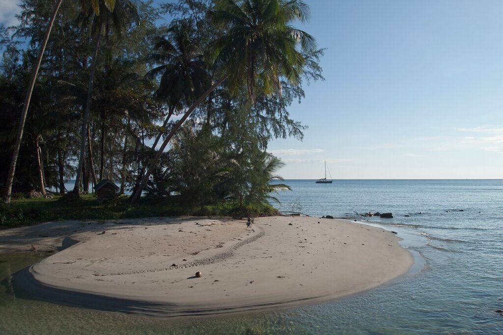 Peaceful sandbank on Koh Kood, Thailand, with palm trees, calm sea, and a sailboat in the distance.
