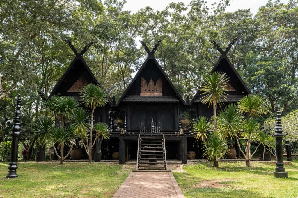 Traditional dark teakwood buildings at the Black House (Baan Dam Museum) in Chiang Rai, Thailand, surrounded by trees and tropical plants.