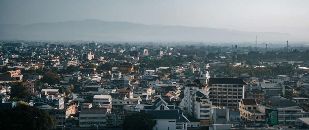 Panoramic view of Chiang Rai city with low-rise buildings, temples, and mountains in the background under a hazy sky.