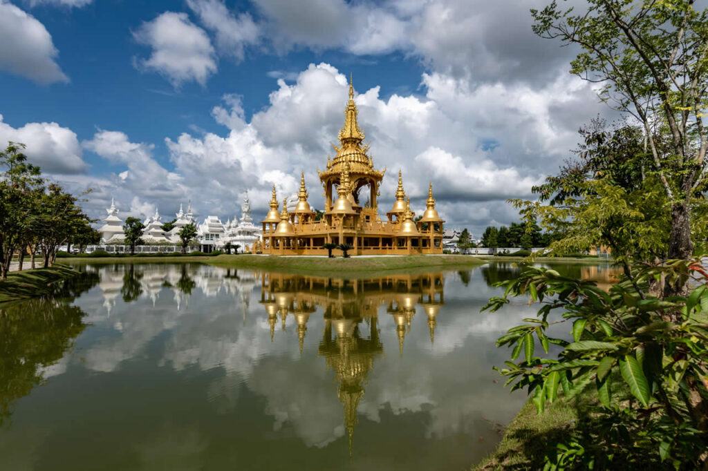 Golden building at Wat Rong Khun (White Temple) complex in Chiang Rai, Thailand, reflected in a pond under a partly cloudy sky.