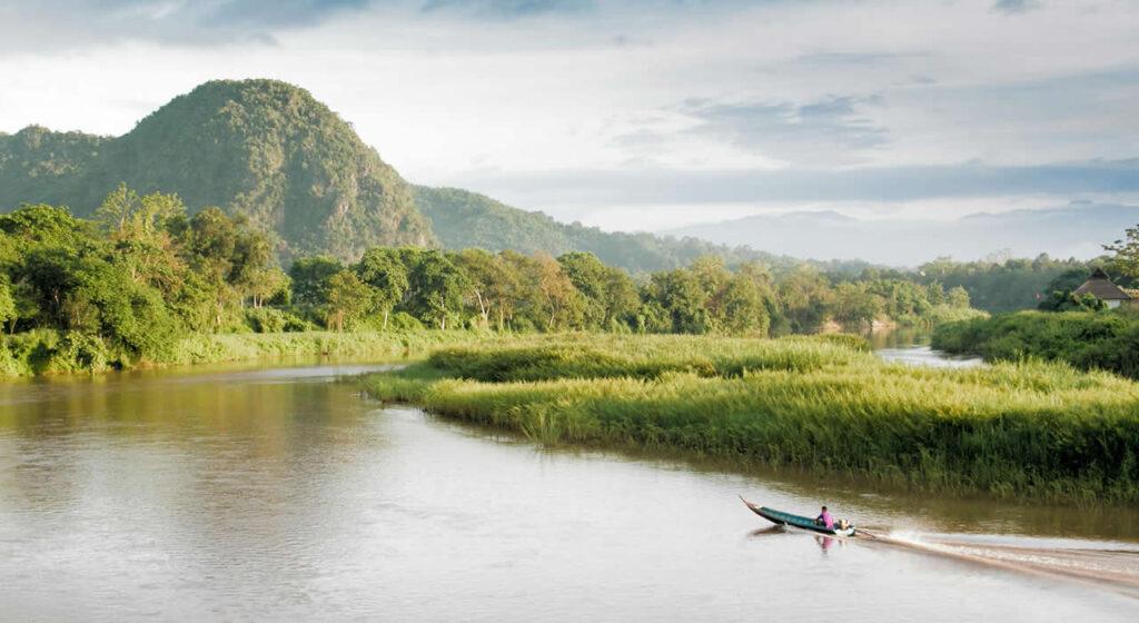 Longtail boat cruising along the Kok River in Chiang Rai, Thailand, with lush green mountains and forests in the background.