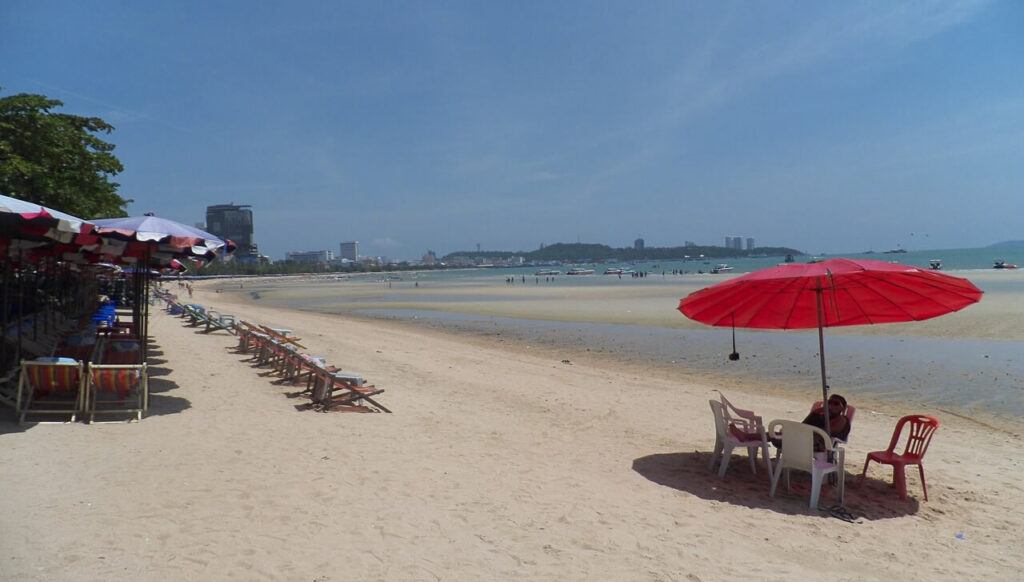 Rows of sunbeds and a red umbrella on Pattaya Beach with city views in the background.