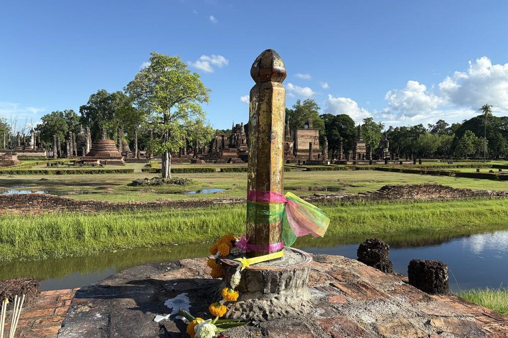 The old city pillar of Sukhothai wrapped in colorful ribbons with ancient temple ruins in the background at Sukhothai Historical Park, Thailand