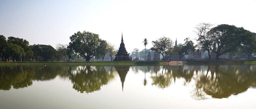 Peaceful morning view of a lake with temple stupas and trees reflected in the water at Sukhothai Historical Park, Thailand