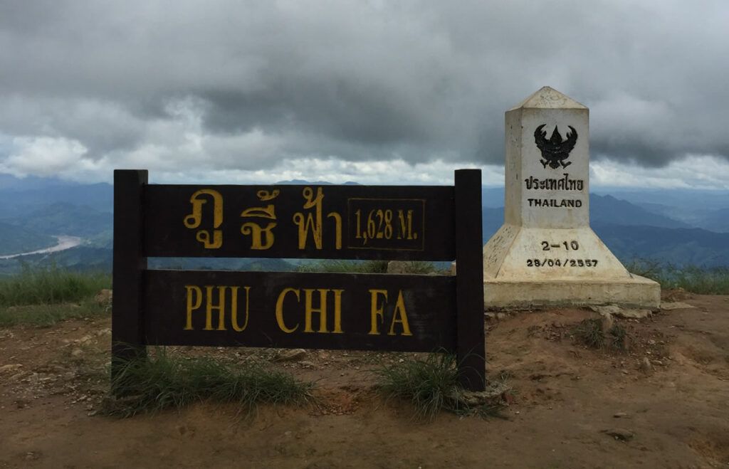 Phu Chi Fa summit sign and Thailand border marker at 1,628 meters, overlooking mountain scenery in Chiang Rai.