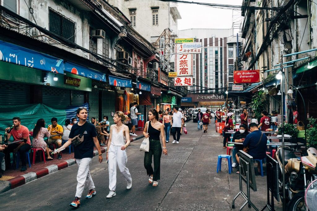 People walking through a busy Bangkok street lined with local food stalls, shopfronts, and overhead cables