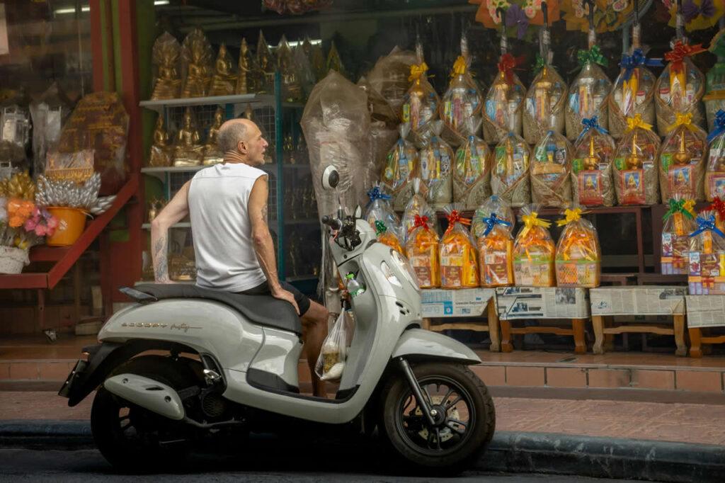 Man sitting on a parked scooter in front of a Thai shop selling religious offerings and golden Buddha statues