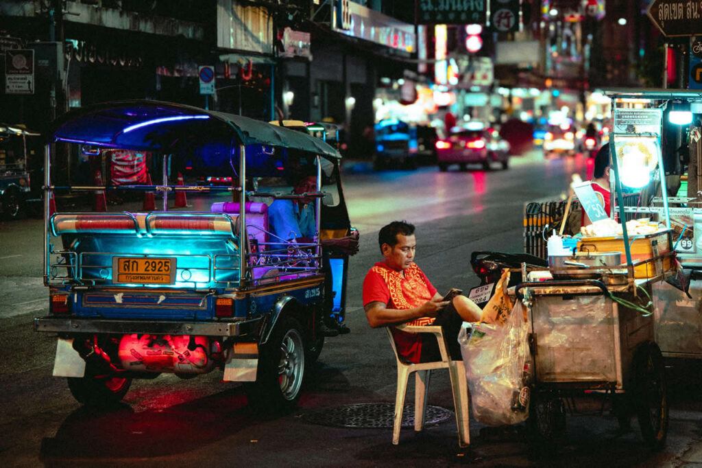 Tuk-tuk parked beside a street food vendor at night on a busy Bangkok road with neon lights and passing traffic