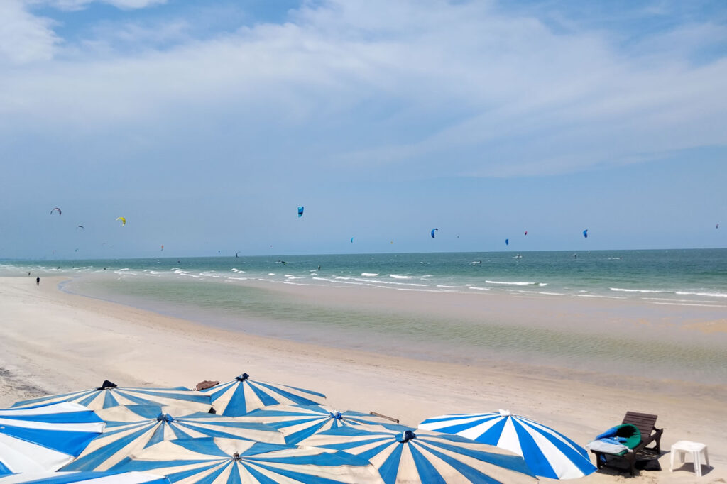 Kiteboarders along the wide sandy beach in Hua Hin with blue beach umbrellas and calm coastal scenery in Thailand.