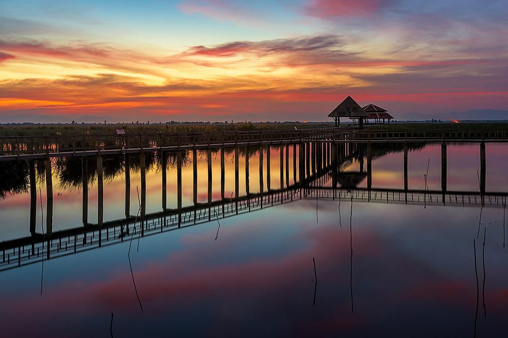 Wooden boardwalk and roofed shelters reflected in the calm water of Bueng Bua wetlands beneath an orange and pink sunset sky.