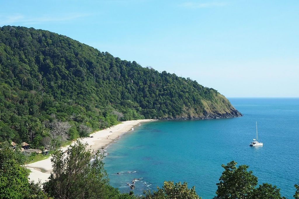 Sandy bay on Koh Lanta bordered by a forested hillside, with turquoise water and a sailboat offshore.