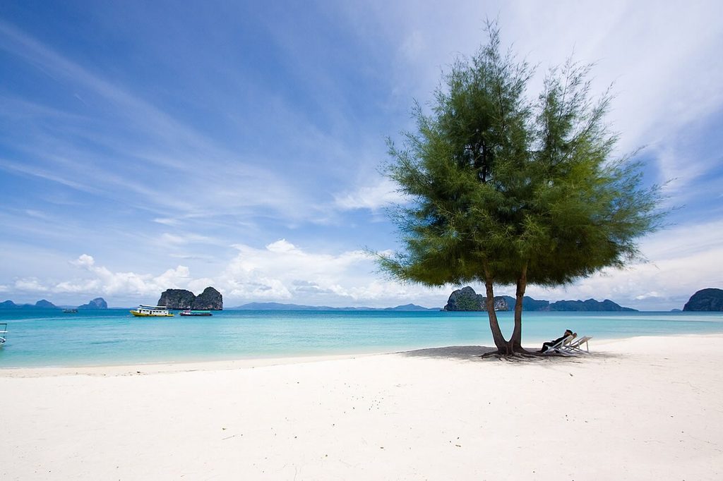 White sandy beach on Koh Ngai with a shady tree, turquoise water, boats and limestone islands offshore.
