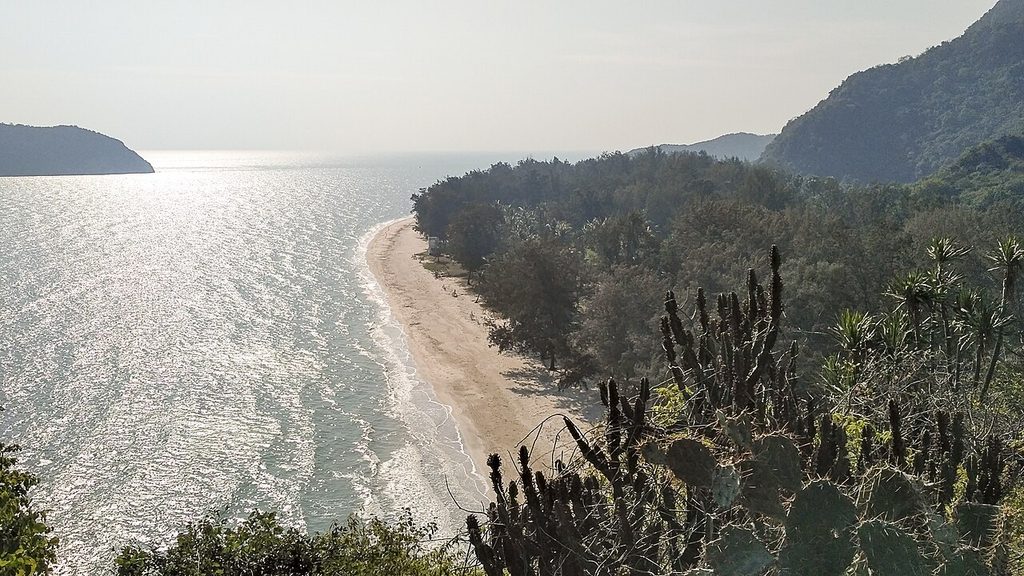 View from a hillside over Laem Sala Beach, with a curved sandy shoreline, sunlit sea and forested mountains.