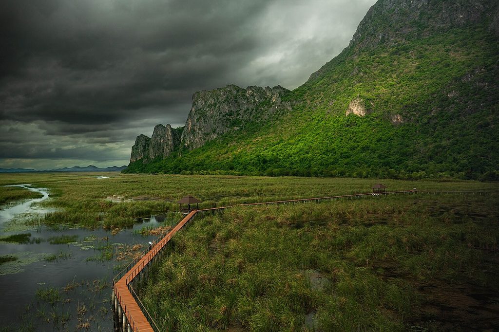 Dark storm clouds above limestone mountains and a wooden boardwalk crossing wetlands in Khao Sam Roi Yot National Park.